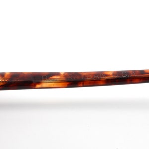 May include: Close-up of a portion of eyeglasses with a tortoiseshell pattern. The text "B. RAY-BAN U.S.A." is visible on the arm of the glasses. The glasses have a classic, retro style.