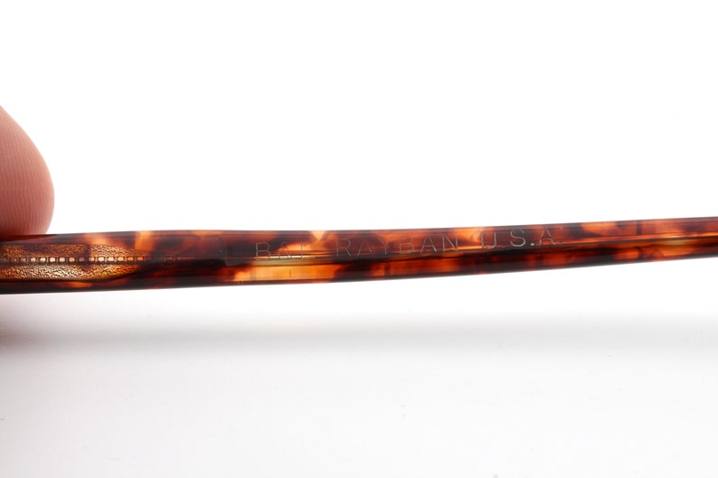 May include: Close-up of a portion of eyeglasses with a tortoiseshell pattern. The text "B. RAY-BAN U.S.A." is visible on the arm of the glasses. The glasses have a classic, retro style.