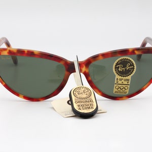 May include: Tortoiseshell Ray-Ban sunglasses with green lenses. The sunglasses have a label that says "Ray-Ban 6-15" Lens XLT" and a sticker with the Olympic rings. The sunglasses are accompanied by a black tag that says "Original Bausch & Lomb."