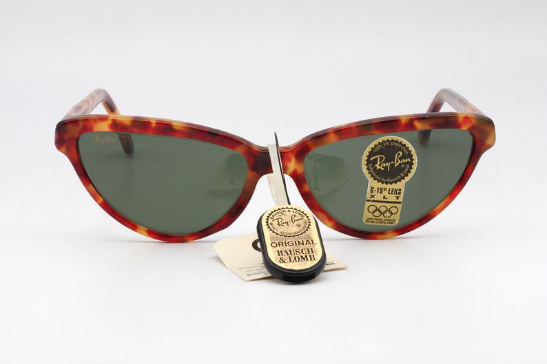May include: Tortoiseshell Ray-Ban sunglasses with green lenses. The sunglasses have a label that says "Ray-Ban 6-15" Lens XLT" and a sticker with the Olympic rings. The sunglasses are accompanied by a black tag that says "Original Bausch & Lomb."