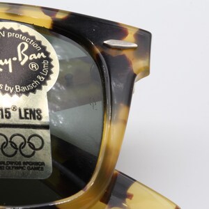 Puede incluir: Primer plano de gafas de sol Ray-Ban con montura carey. Las gafas de sol tienen una pegatina que dice "100% UV protection" y "G-15 Lens". La pegatina tambi&eacute;n menciona "Sunglasses by Bausch & Lomb" y "1962 Olympic Games".