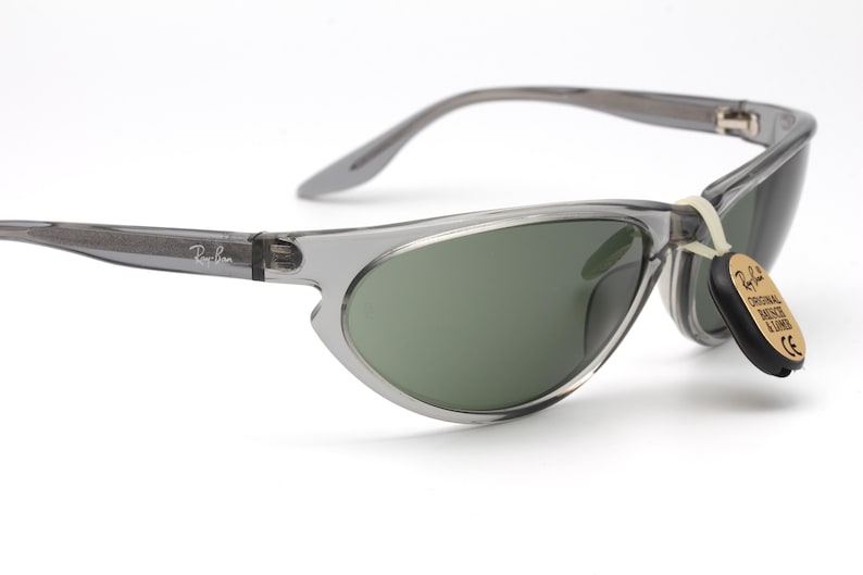May include: Gray cat-eye sunglasses with green lenses and a Ray-Ban tag attached. The sunglasses have a clear frame.