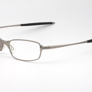 May include: A pair of eyeglasses with a silver metal frame and rectangular lenses. The temples have black rubberized tips. The glasses are set against a white background.