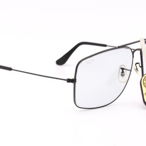 May include: Black Ray-Ban square-shaped glasses with clear lenses. The glasses have a thin metal frame and a small metal nose bridge. The glasses are sitting on a white surface. A small tag with the text "ORIGINAL BAUSCH & LOMB" is attached to the glasses.