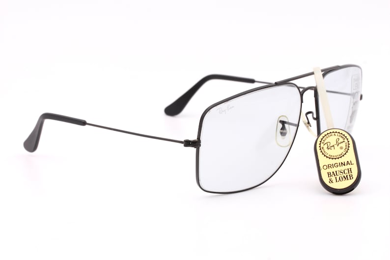 May include: Black Ray-Ban square-shaped glasses with clear lenses. The glasses have a thin metal frame and a small metal nose bridge. The glasses are sitting on a white surface. A small tag with the text "ORIGINAL BAUSCH & LOMB" is attached to the glasses.