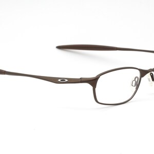 May include: Brown metal frame spectacles with clear lenses. The frame has a thin, sleek design and features a small, circular logo on the side.