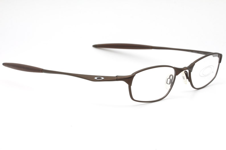 May include: Brown metal frame spectacles with clear lenses. The frame has a thin, sleek design and features a small, circular logo on the side.