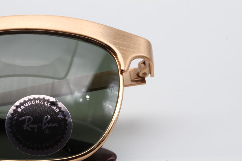 May include: Gold-tone metal frame sunglasses with green lenses. The sunglasses have a Ray-Ban logo on the left lens.