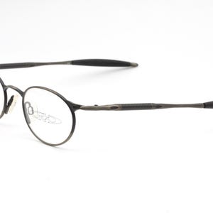 May include: A pair of round, silver-toned eyeglasses with thin metal frames. The arms of the glasses are black and have a rubberized texture.