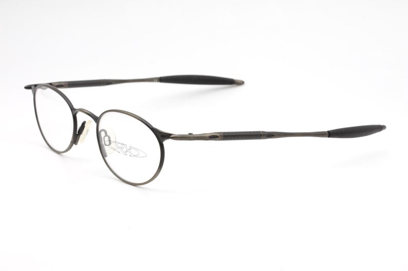 May include: A pair of round, silver-toned eyeglasses with thin metal frames. The arms of the glasses are black and have a rubberized texture.