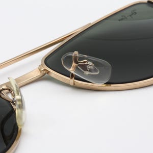 May include: Close-up of gold-framed sunglasses with dark lenses. The sunglasses have clear nose pads and a gold bridge. The lenses are a dark green color. The frame is a classic aviator style.