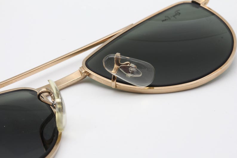 May include: Close-up of gold-framed sunglasses with dark lenses. The sunglasses have clear nose pads and a gold bridge. The lenses are a dark green color. The frame is a classic aviator style.