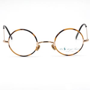 May include: Round tortoiseshell eyeglasses with gold metal frames. The frames have a subtle tortoise pattern and the lenses are clear. The brand name "The Ralph Lauren" is printed on the right lens.