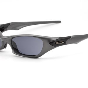 Może przedstawiać: Szare okulary przeciwsłoneczne z ciemnymi soczewkami. Okulary mają zakrzywioną oprawkę i elegancki, nowoczesny design. Logo Oakley jest widoczne na zausznikach. Okulary są na białym tle.