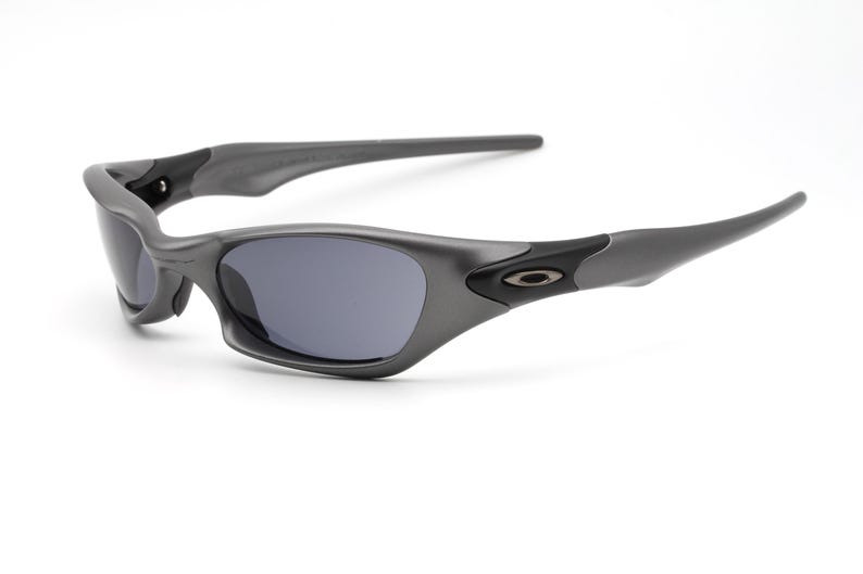 Może przedstawiać: Szare okulary przeciwsłoneczne z ciemnymi soczewkami. Okulary mają zakrzywioną oprawkę i elegancki, nowoczesny design. Logo Oakley jest widoczne na zausznikach. Okulary są na białym tle.