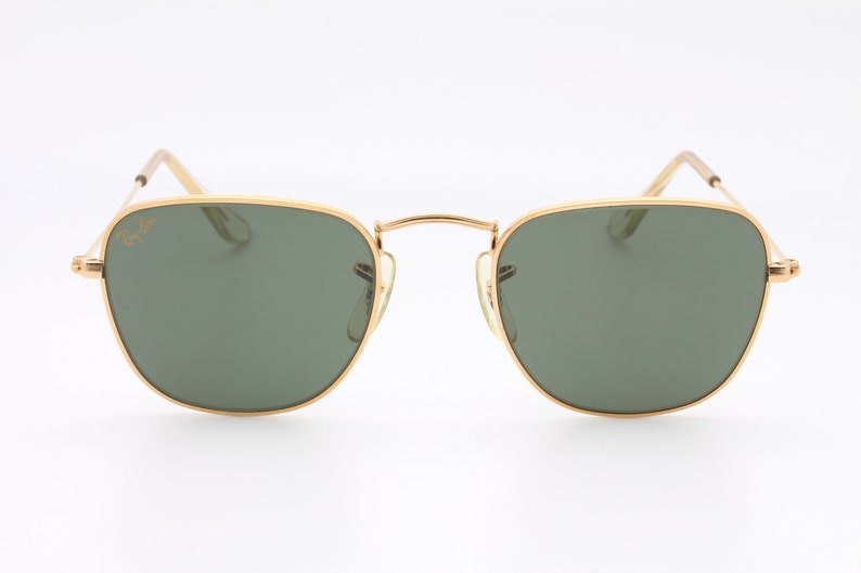 May include: Gold-framed sunglasses with green lenses. The sunglasses have a classic, rectangular shape.