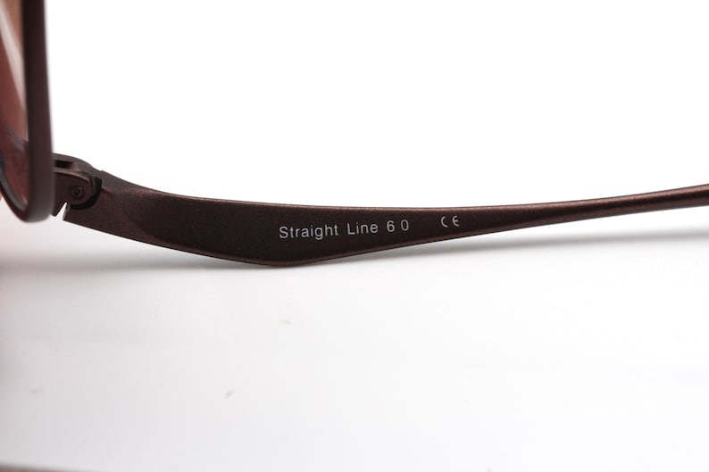 May include: Close-up of the brown metal arm of a pair of glasses with the text "Straight Line 6.0 CE" printed on it.