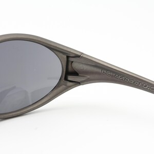 May include: Close-up of gray sunglasses with dark lenses. The frame is a matte gray color. The text "MADE IN U.S.A." is printed on the arm of the sunglasses.