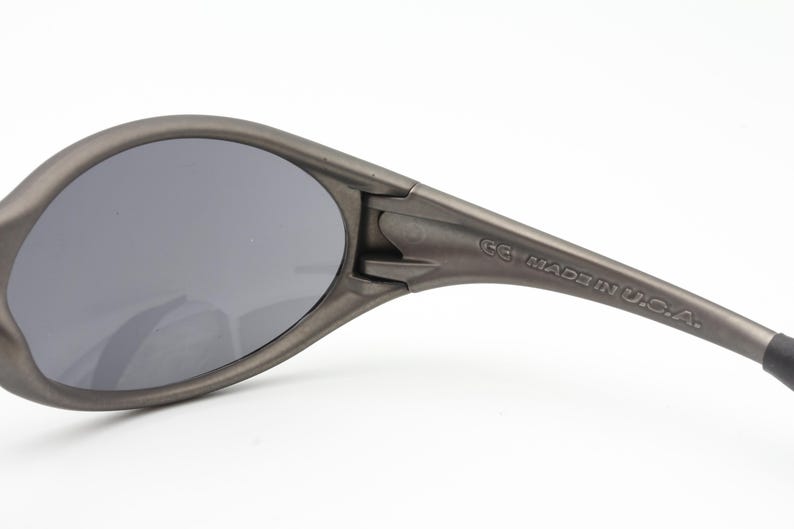 May include: Close-up of gray sunglasses with dark lenses. The frame is a matte gray color. The text "MADE IN U.S.A." is printed on the arm of the sunglasses.