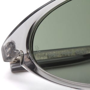 May include: Close-up of a pair of silver-toned sunglasses with green lenses. The sunglasses have a thin, oval frame and a subtle inscription on the lens that reads "IB".