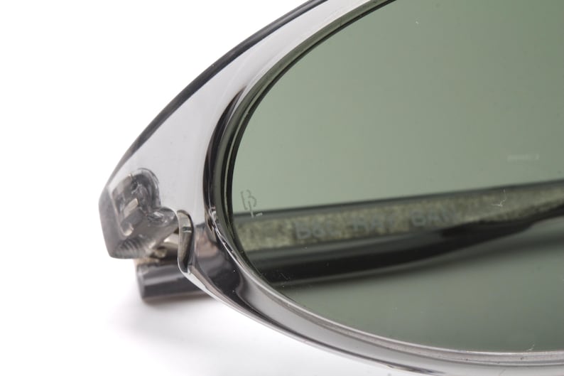 May include: Close-up of a pair of silver-toned sunglasses with green lenses. The sunglasses have a thin, oval frame and a subtle inscription on the lens that reads "IB".