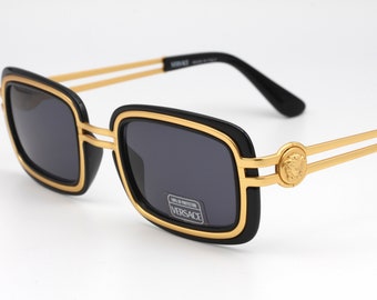Puede incluir: Gafas de sol cuadradas negras y doradas con un logotipo dorado de Versace en el lateral. Las gafas tienen una montura negra con un borde dorado.