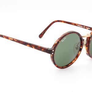 May include: Tortoiseshell Ray-Ban sunglasses with green lenses and gold accents. The sunglasses have a round frame and the text "Ray-Ban" is visible on the right lens. The text "Premier C-1-W-595" is visible on the right arm.