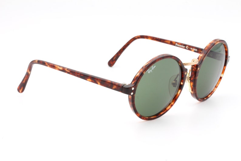 May include: Tortoiseshell Ray-Ban sunglasses with green lenses and gold accents. The sunglasses have a round frame and the text "Ray-Ban" is visible on the right lens. The text "Premier C-1-W-595" is visible on the right arm.