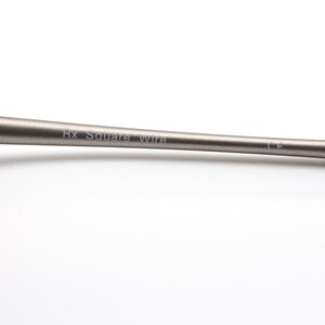 May include: Close-up of a metallic gray Rx Square Wire. The text is printed in a thin, sans-serif font. The wire has a smooth, matte finish and tapers slightly towards the ends. The background is plain white.