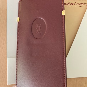 May include: A burgundy leather wallet with gold accents and a Cartier logo. The wallet has a stitched border and is closed.