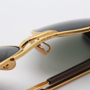 May include: Close-up of gold-tone Ray-Ban sunglasses with dark green lenses. The sunglasses are folded in half, showing the Ray-Ban branding on the frame.
