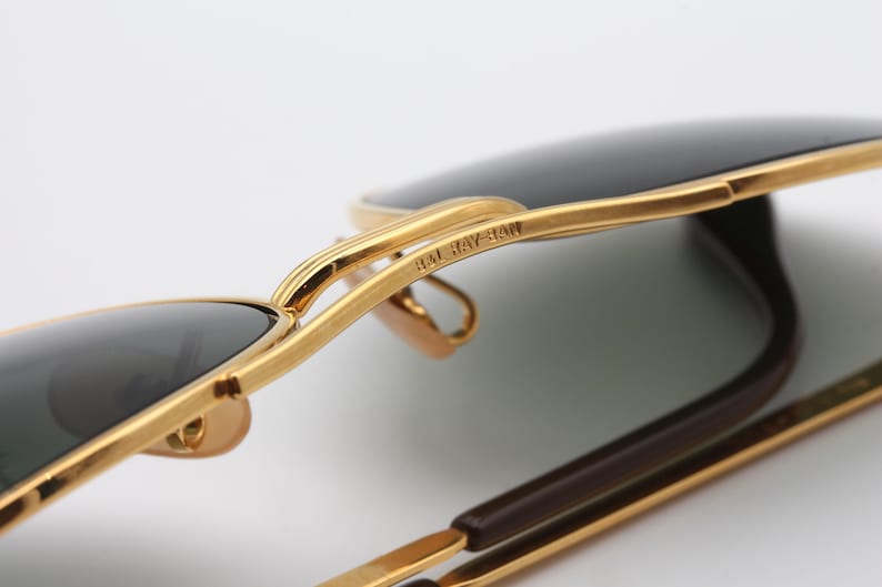 May include: Close-up of gold-tone Ray-Ban sunglasses with dark green lenses. The sunglasses are folded in half, showing the Ray-Ban branding on the frame.