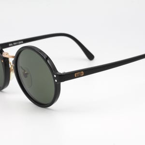 May include: Black round sunglasses with green lenses and gold accents. The sunglasses have the text "Ray-Ban" on the side.