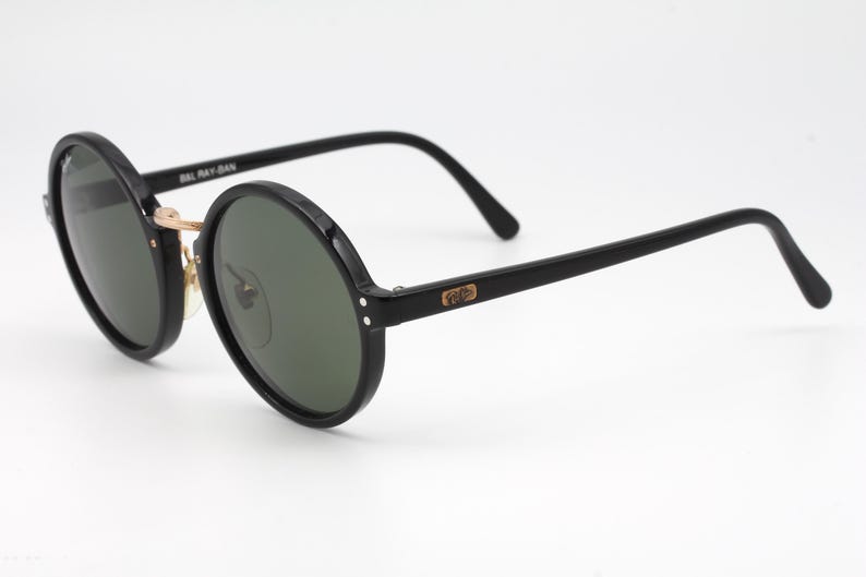 May include: Black round sunglasses with green lenses and gold accents. The sunglasses have the text "Ray-Ban" on the side.