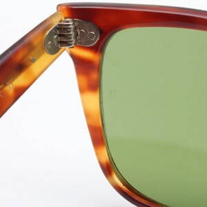 May include: Close-up of vintage sunglasses with a tortoiseshell pattern frame and green lenses. The hinge is visible, and the arm of the glasses has the word "ZAREER" printed on it. The sunglasses are set against a white background.