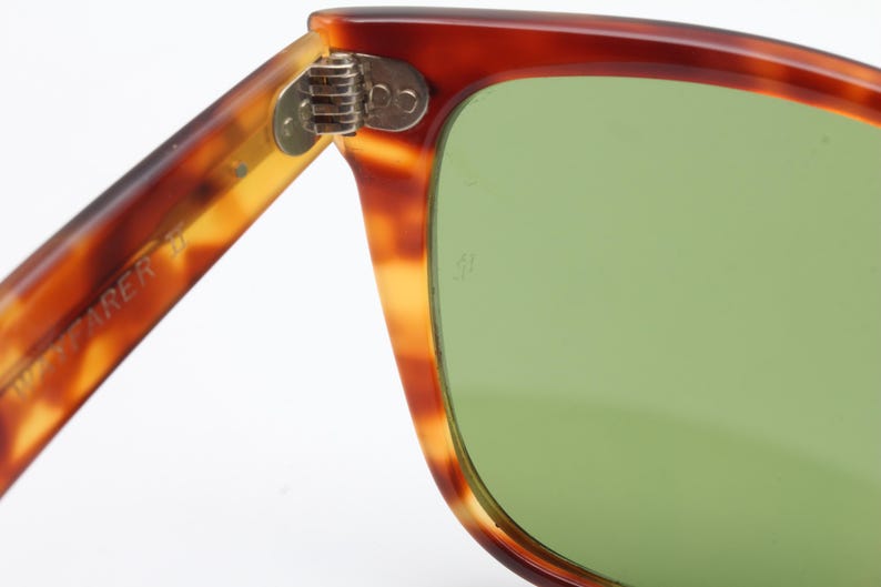 May include: Close-up of vintage sunglasses with a tortoiseshell pattern frame and green lenses. The hinge is visible, and the arm of the glasses has the word "ZAREER" printed on it. The sunglasses are set against a white background.