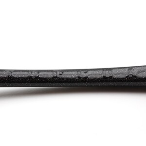 Puede incluir: Primer plano de un marco de raqueta de tenis negro con la marca "HEAD" visible. El marco tiene una textura moteada y un dise&ntilde;o curvo, con un agarre negro en el extremo. El fondo es blanco liso.