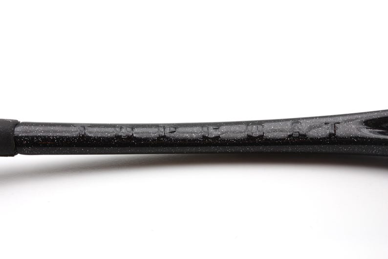 Puede incluir: Primer plano de un marco de raqueta de tenis negro con la marca "HEAD" visible. El marco tiene una textura moteada y un dise&ntilde;o curvo, con un agarre negro en el extremo. El fondo es blanco liso.