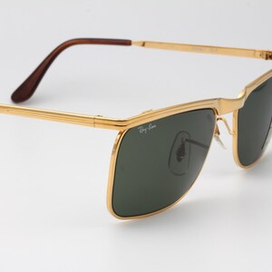 Op de afbeelding: Goudkleurige zonnebril met donkergroene glazen. De zonnebril heeft een rechthoekige vorm en bruine pootjes. Het Ray-Ban-logo is zichtbaar op het glas. De bril staat tegen een witte achtergrond.