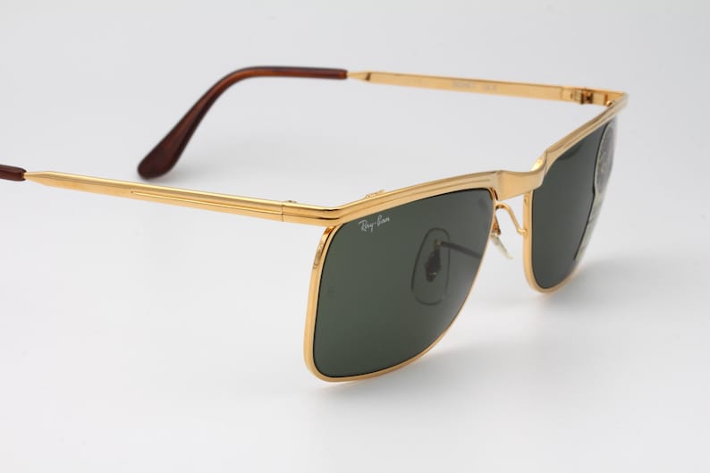 Op de afbeelding: Goudkleurige zonnebril met donkergroene glazen. De zonnebril heeft een rechthoekige vorm en bruine pootjes. Het Ray-Ban-logo is zichtbaar op het glas. De bril staat tegen een witte achtergrond.