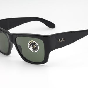 May include: Black Ray-Ban sunglasses with dark green lenses. The sunglasses have a thick, rectangular frame and the Ray-Ban logo is visible on the temple. A sticker is on the lens.