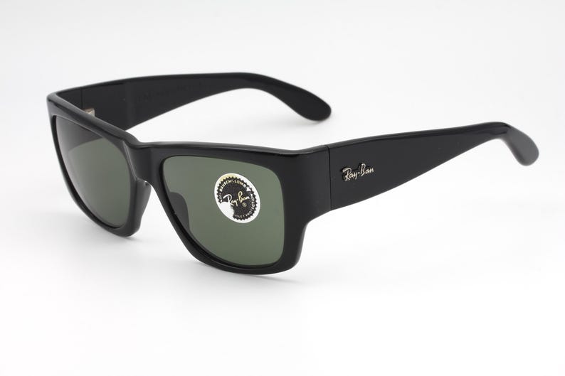 May include: Black Ray-Ban sunglasses with dark green lenses. The sunglasses have a thick, rectangular frame and the Ray-Ban logo is visible on the temple. A sticker is on the lens.