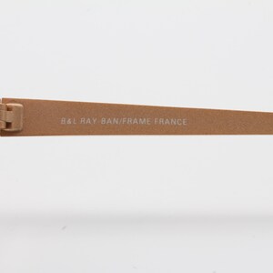 May include: Close-up of the gold-toned metal arm of a pair of Ray-Ban sunglasses. The inscription "BAL RAY-BAN/FRAME FRANCE" is visible on the arm.
