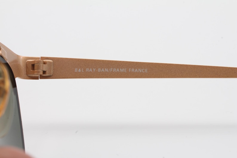 May include: Close-up of the gold-toned metal arm of a pair of Ray-Ban sunglasses. The inscription "BAL RAY-BAN/FRAME FRANCE" is visible on the arm.
