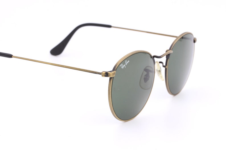 Puede incluir: Gafas de sol redondas doradas con lentes verdes y el logotipo de Ray-Ban en el marco.