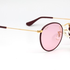 May include: Round pink sunglasses with a gold frame and burgundy accents. The sunglasses are on a white background.