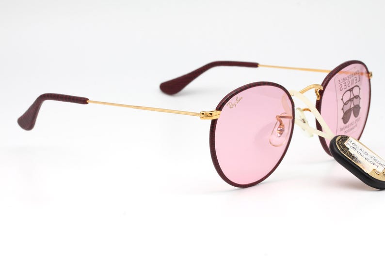 May include: Round pink sunglasses with a gold frame and burgundy accents. The sunglasses are on a white background.