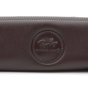 Może przedstawiać: Brązowe sk&oacute;rzane etui na okulary przeciwsłoneczne Ray-Ban z okrągłym logo z napisem "Ray-Ban" i "Genuine Leather".