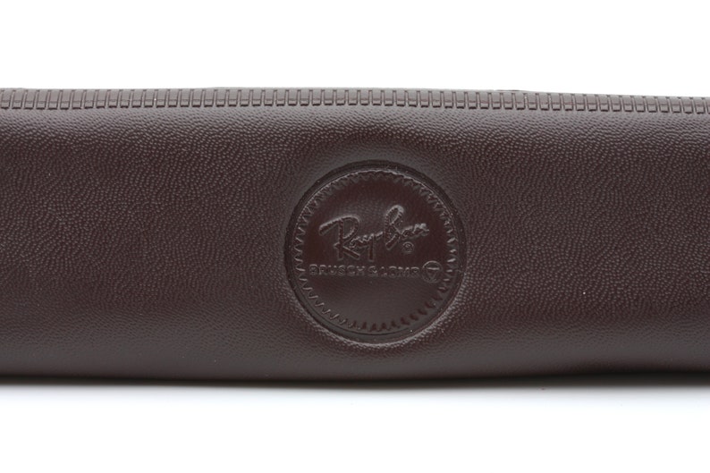 Może przedstawiać: Brązowe sk&oacute;rzane etui na okulary przeciwsłoneczne Ray-Ban z okrągłym logo z napisem "Ray-Ban" i "Genuine Leather".