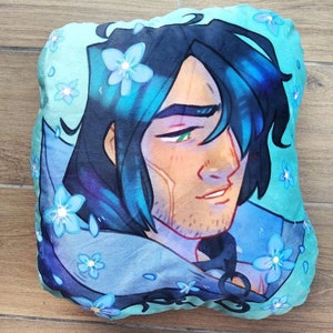 May include: A blue and green pillow with a cartoon illustration of a man with blue hair and a light blue scarf. The man has a light brown complexion and green eyes. The background is a light blue with small white flowers.